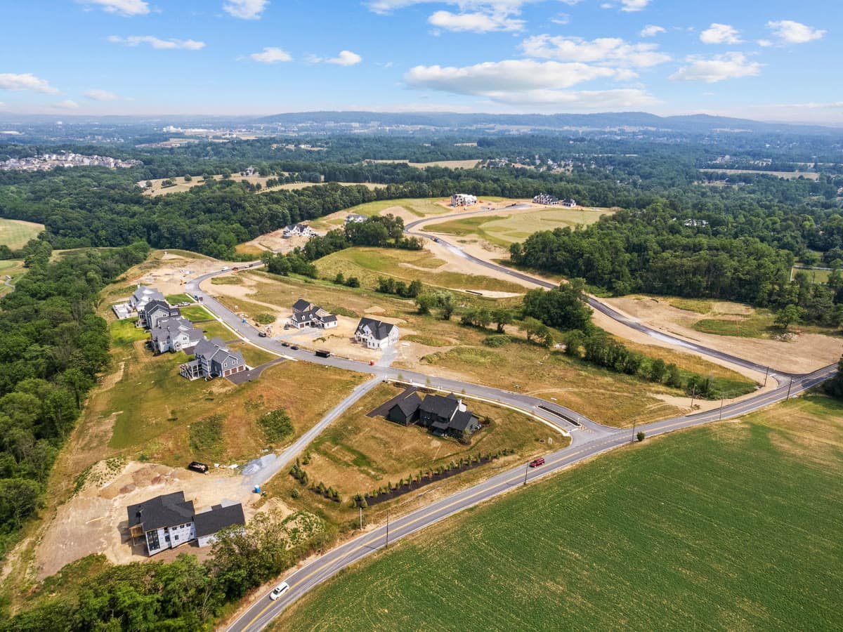 An aerial view of new homes going up along curving roads, with farmland and wooded hills around them