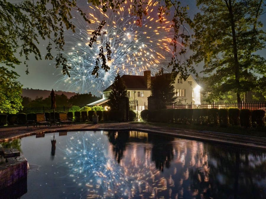 Fireworks over a white house at night, reflected in a pool