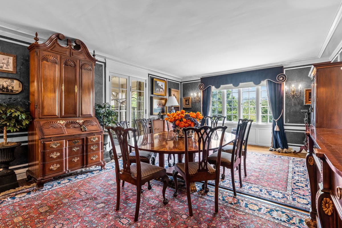 A dining room with an antique secretary, a round table set with orange flowers, and an oriental rug