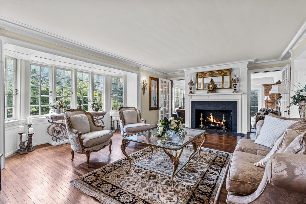 A living room with a lit fire, a glass coffee table, French armchairs, and a bay of windows