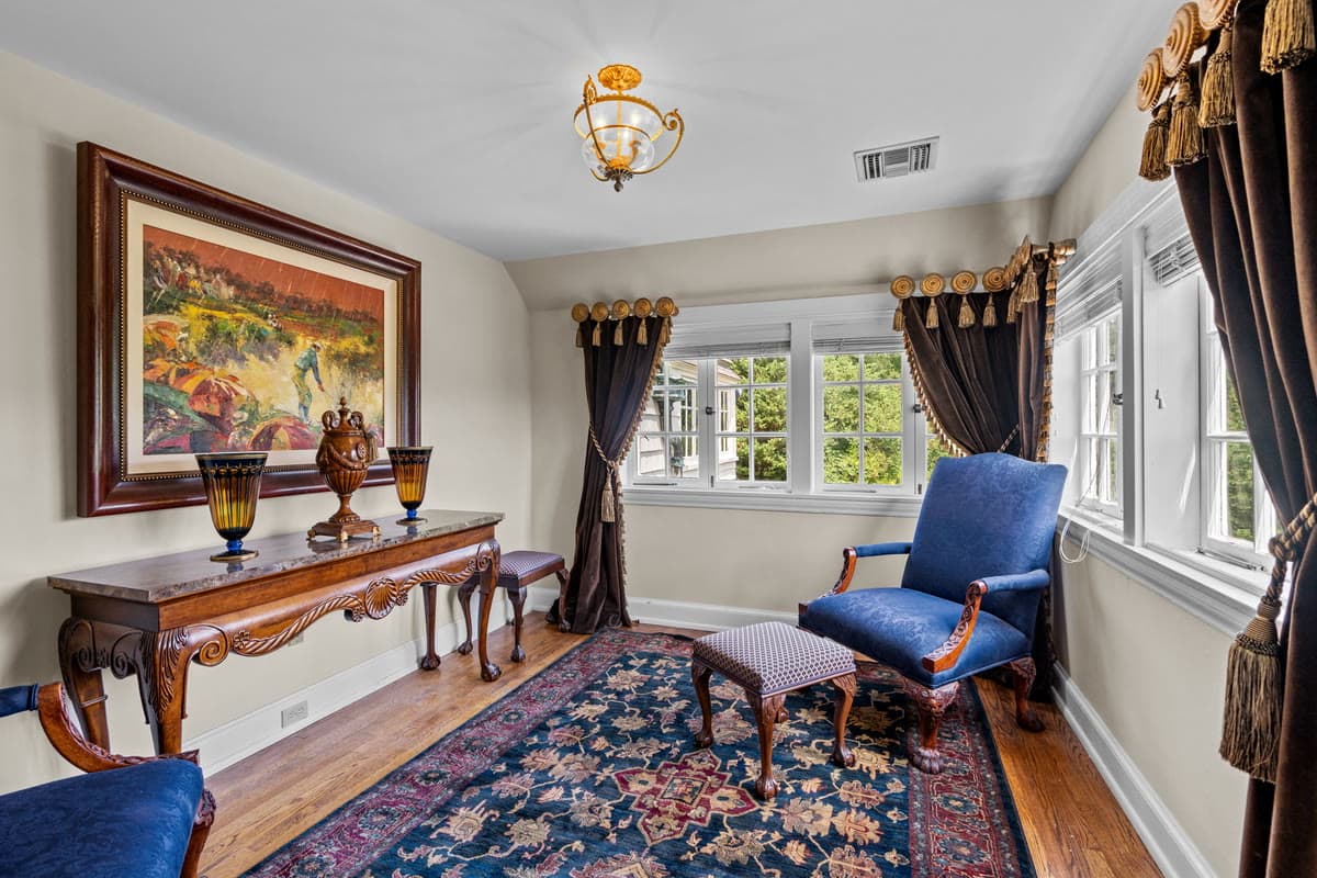 A sitting room with a large landscape painting over a carved console and a blue armchair by the window