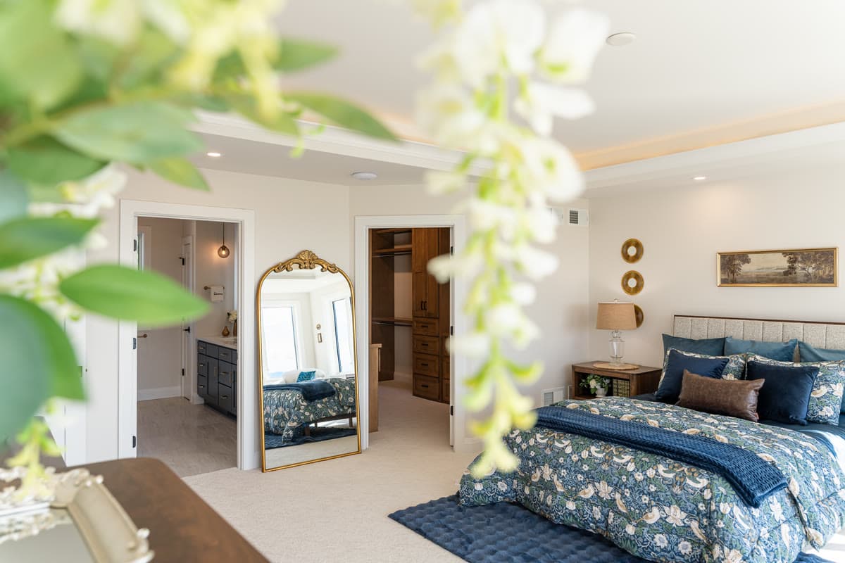 A bedroom with a floral quilt and navy pillows, seen past a spray of white blossom, with a gilded floor mirror by the closet
