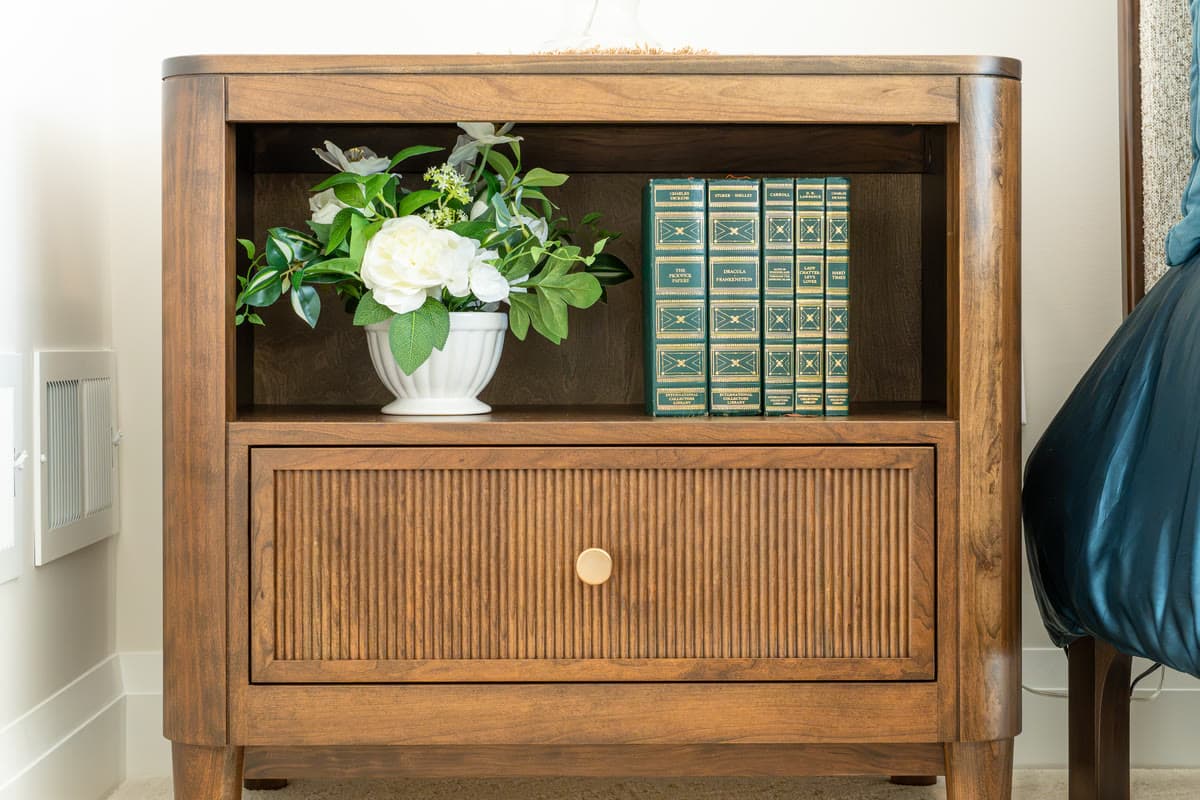 A wooden nightstand with white flowers and a row of green bound books