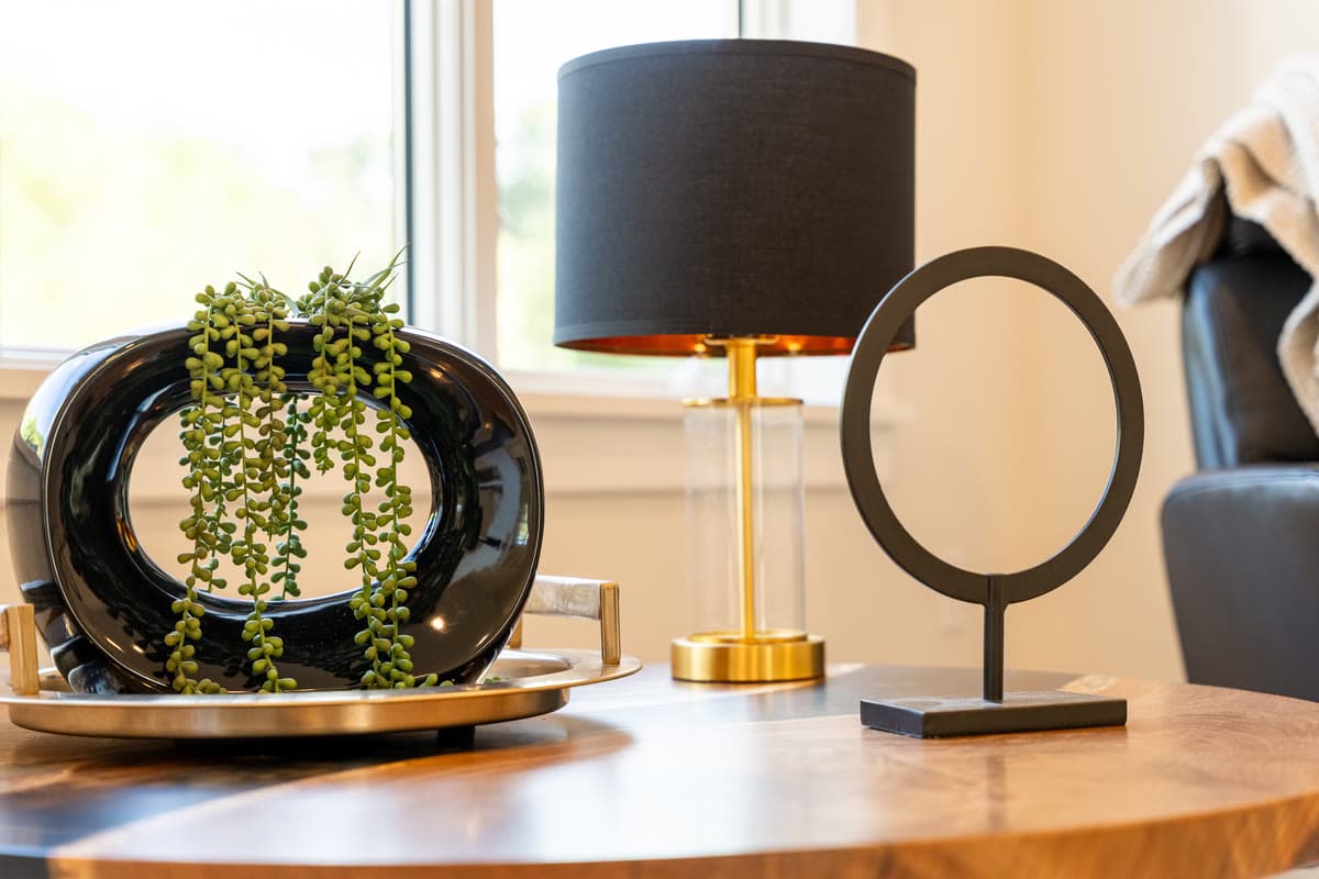A black ring vase with trailing greenery beside a black shaded lamp on a wood side table