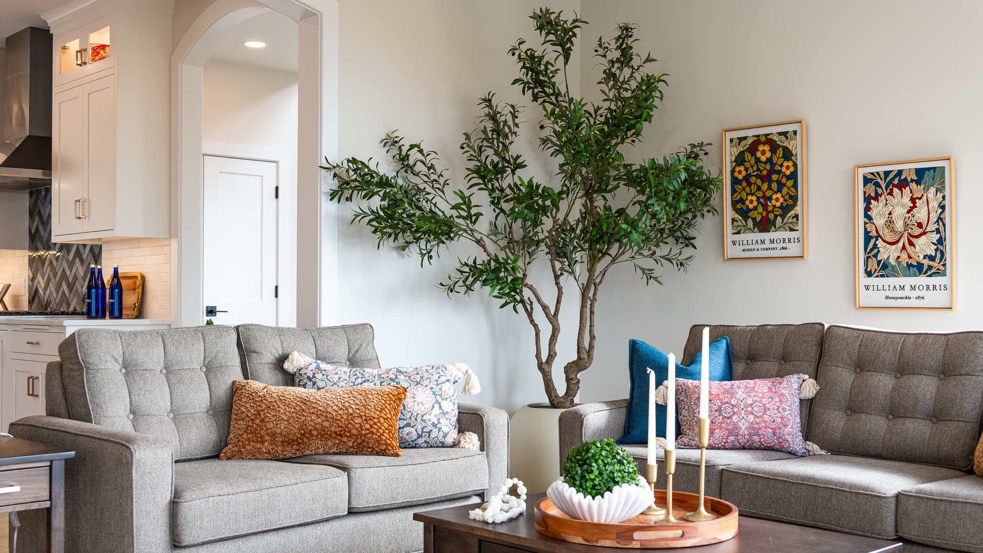A living room with two grey tufted sofas, a potted olive tree, and William Morris prints on the wall