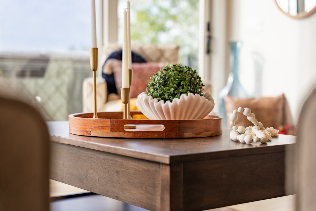 A wooden tray with brass candlesticks and a fluted white bowl of greenery on a dining table