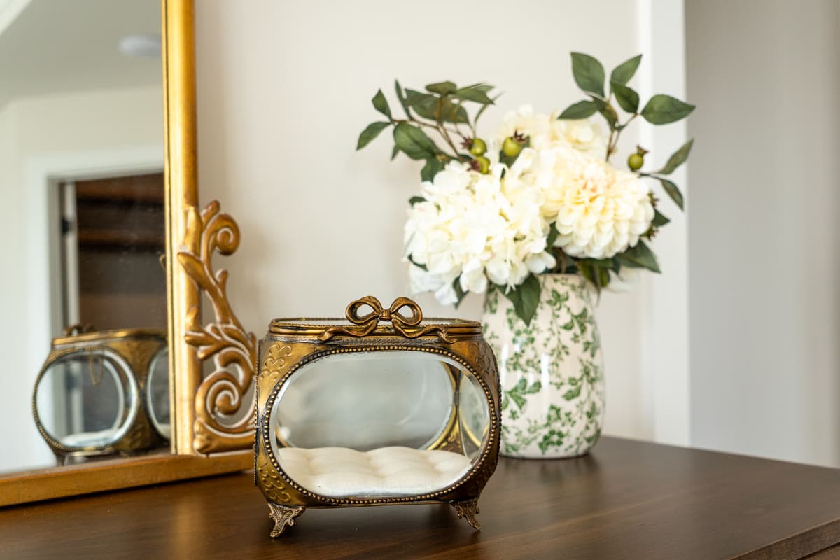 A gilded mirror, a brass and glass jewelry box, and white flowers in a green and white vase
