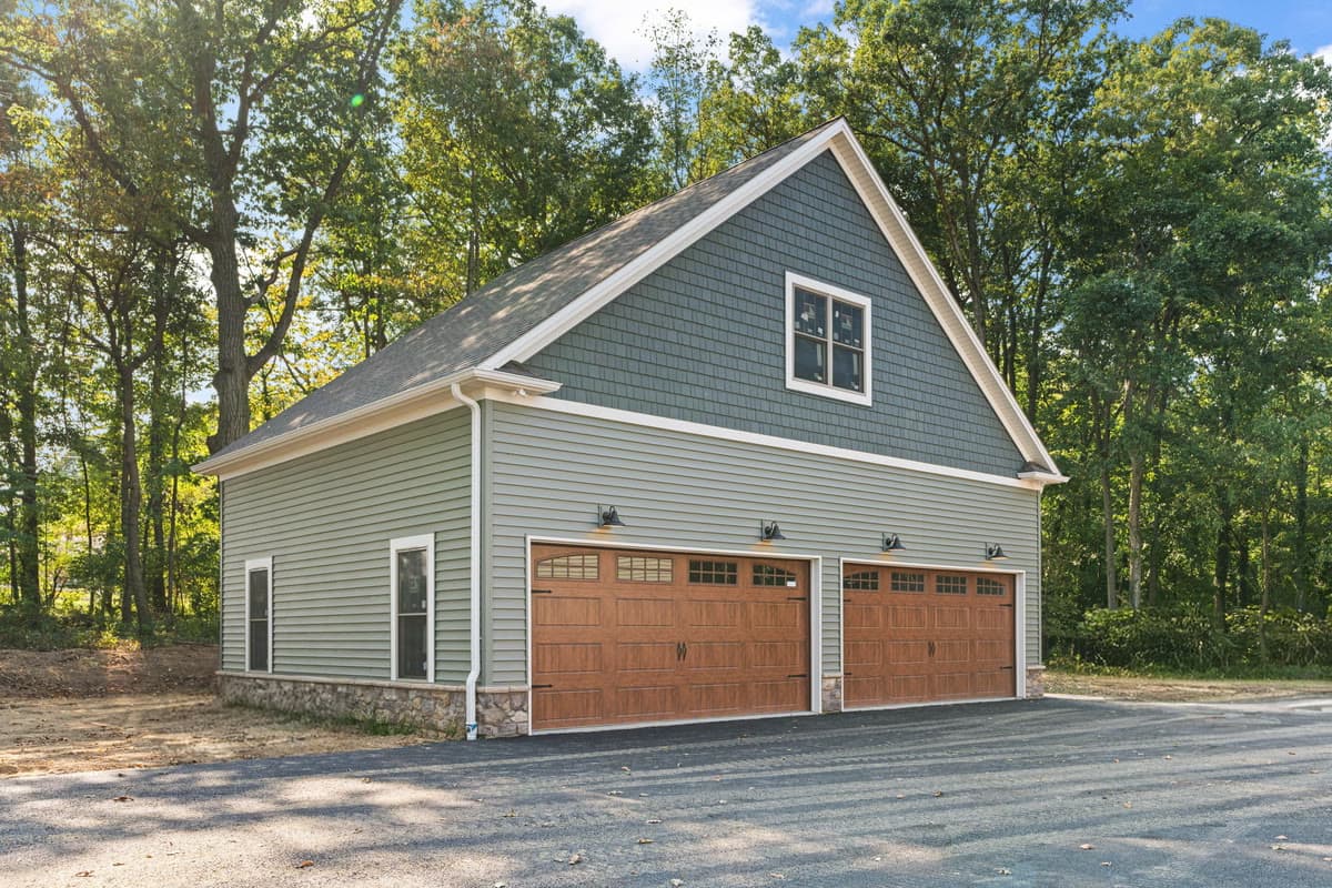 A detached three bay garage in green siding with wood doors, at the edge of woodland