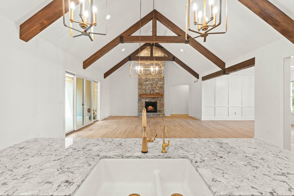 A view across a marble island to a vaulted great room with timber trusses and a stone fireplace